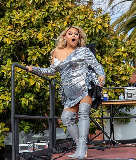 Jiggly Caliente, wearing a sparkly silver dress, thigh-high boots, and fishnet stockings, performs on an outdoor stage.