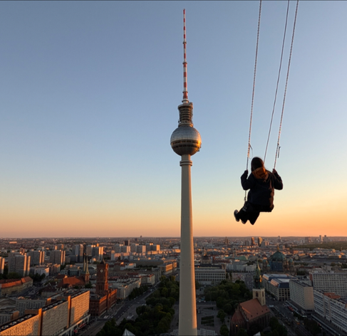 Das Bild zeigt eine Person, die auf einer Schaukel über dem Fernsehturm in Berlin schwingt. Der Fernsehturm, ein markantes Wahrzeichen Berlins, ragt majestätisch in den Himmel und ist im Vordergrund des Bildes deutlich zu sehen. Die Person, die auf der Schaukel sitzt, ist gegen das Licht des Abendhimmels abgesetzt und schwingt vor dem Hintergrund der Stadt. Die Stadt Berlin erstreckt sich weit in die Ferne, mit einer Vielzahl von Gebäuden, die von der Sonne des Abendhimmels in warmes Licht getaucht werden. Der Himmel ist klar und zeigt eine sanfte Farbverläufe von Blau zu Orange, was auf die Dämmerung hinweist. Die Schaukel befindet sich auf einer Höhe, die den Fernsehturm deutlich übersteigt, was eine beeindruckende Perspektive bietet.