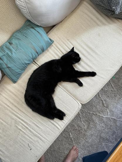 A black cat is in a moon shape. Lying on the beige couch with pillows of various shapes.