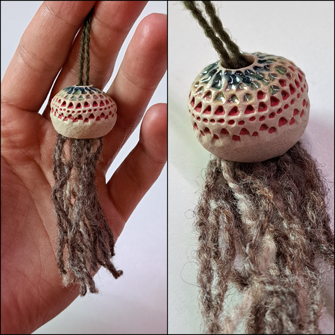A tassel with it's cap made out of stoneware clay with small holes in it. The holes are filled with red engobe lower down and green and blue glaze at the top. The temporary tassel itself is made out of grey yarn and is quite fluffy and thin.