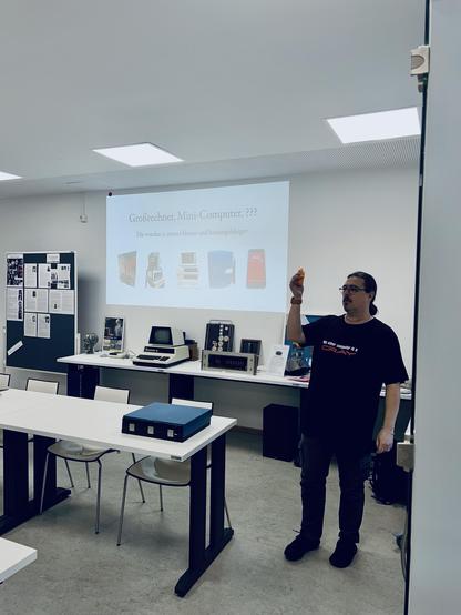 An individual is giving a presentation in a classroom-like setting. A projector screen displays images of various computers labeled “Großrechner, Mini-Computer, ???” indicating a discussion about the evolution of computers. The presenter, holding an orange object,