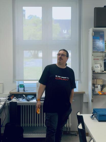 A person with long hair and glasses stands in an indoor space, wearing a black t-shirt that says "My other computer is a CRAY." They are near a table with various electronic devices and tools, with large windows in the background.