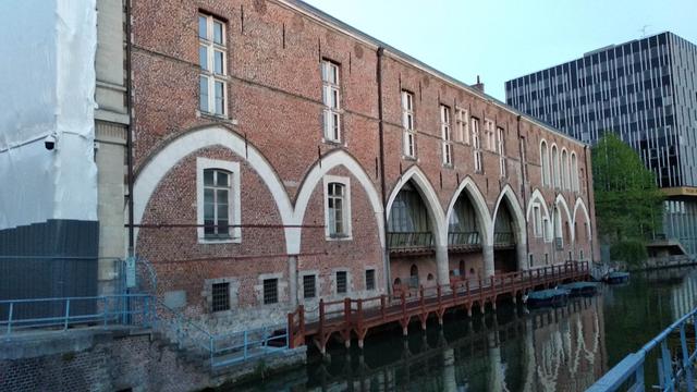 Un bâtiment en briques avec de belles arches blanches dans la partie basse et des hautes fenêtres au premier étage, au bord de la Scarpe, avec des barques attachées le long du bâtiment. En arrière plan, un bâtiment moderne moche