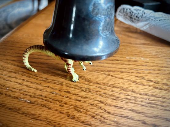 A green Dilophosaurus with brown spots stands on a tan, wooden table with a dirty silver bell over their head. A white, wrinkled doily is in the background.
