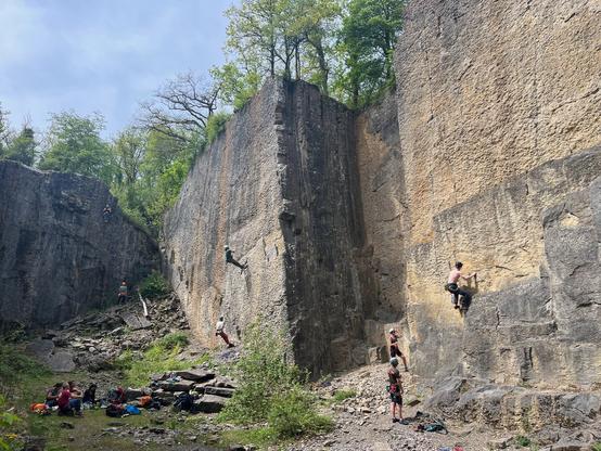 A small climbing crag with multiple climbers active on the wall. A small group of people is sitting in the grass at the base of the rock face, watching the climbers.