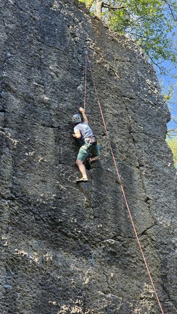 A climber on the wall, locking off his left arm while reaching up towards a small hold. He's protected by a top-rope.