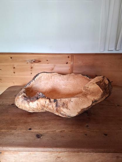 A large, rustic wooden bowl carved from a spruce burl, resting on an aged wooden larch table. The bowl has an irregular, organic shape, with a rough, textured exterior showing bark remnants and natural knots, while the interior is smoothly hollowed out. The setting is cozy with a wooden paneled walls.