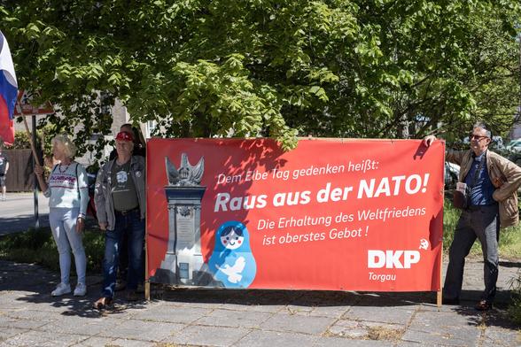 DKP am Torgauer Thälmann-Monument. Auf ihrem Banner steht: "Dem Elbe-Tag gedenken heißt: Raus aus der Nato! Die Erhaltung des Weltfriedens ist oberstes Gebot!" Darauf ist eine Matruschka-Puppe mit einer Friedenstaube darauf abgebildet.