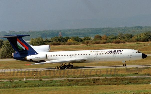 Side view of a white, 3 engined jet airliner with the engines mounted on the rear fuselage, taxiing from left to right.
The plane is mostly white, with blue "Malev" titles on the upper forward fuselage, with smaller "Hungarian Airlines" titles and a small Hungarian flag just aft of that.
There are small "TU-154" titles on the lower forward fuselage, just aft of the cockpit.
The tail and rear fuselage is dark blue, with green, white, and red diagonal stripes on the tail, vaguely resembling an upper-case letter "M", but actually representing the Hungarian flag.
The engine pods are white, with the registration "HA-LCR" on the side-mounted engines in black.
Green grass fills the foreground, with more grass in the background leading up to trees and bushes in the distance, under grey skies.