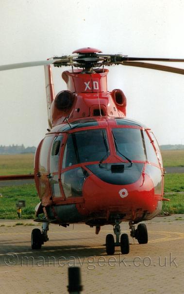 Almost head-on, vertically-oriented front view of a bright red helicopter parked facing slightly off to the right of the camera.
The helo has an extensively glazed cockpit and passenger cabin, with all the bodywork painted red except for the top of the nose cone directly in front of the pilots, and a circular white logo on the nose.
The white upper-case letters "XD" are on the top of the red cowling around the rotor mast .
Grey concrete apron fills the foreground at the bottom of the frame, with green grass leading off to houses in the distance, under hazy sky.