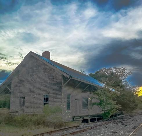 "This image evokes a profound sense of abandonment and loss. It features an old train station, whose wooden structure bears the scars of time and decay. The wood is weathered, chipped, and faded, as though the elements have eroded its former vitality. The roof, clad in rusted metal sheets, hints at a past life filled with activity and purpose. A lone brick chimney stands defiantly atop the building, an echo of warmth that once emanated from within.

The windows, shattered and empty, are like vacant eyes gazing out at the world—once a portal to movement and connection, now filled with silence. The doors remain closed, safeguarding nothing but memories of bygone days. Around the building, nature has taken over; wild bushes, tall grasses, and sprawling trees crowd around the structure as if to reclaim their territory.

Before the station, disused train tracks lie forgotten and forlorn. They are coated in rust, littered with debris, and veiled by the encroaching weeds. The tracks seem to stretch into oblivion, symbolizing journeys that will never resume.

Above it all, the sky mirrors the mood of the station, with dark, brooding clouds mingling with streaks of light. The interplay of shadows and sun illuminates the station in a haunting glow, highlighting its desolation. A yellow tint from the setting sun adds a wistful touch, as though bidding farewell to this piece of history." - Copilot