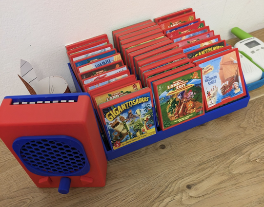 Photo of the audio player. It looks like a Gameboy, but with a speaker instead of a screen. Next to it is a tray with cartridges containing an audio book each. The cartridges are also Gameboy inspired.