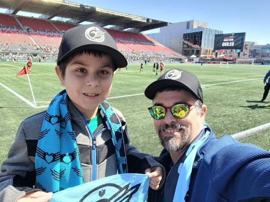 My eldest son & I smiling near the corner flag, wearing teal Rapid FC scarves and a black & white Rapid FC hats.