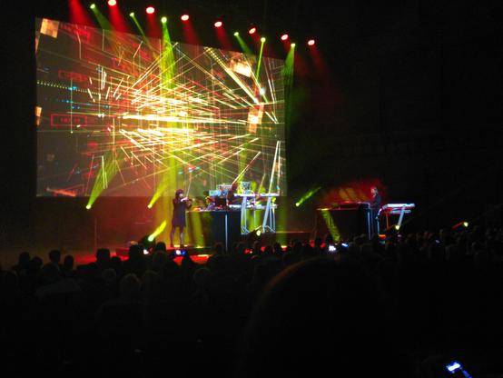 Tangerine Dream performing. The stage is lit in red and green with colorful projections on the backwall.