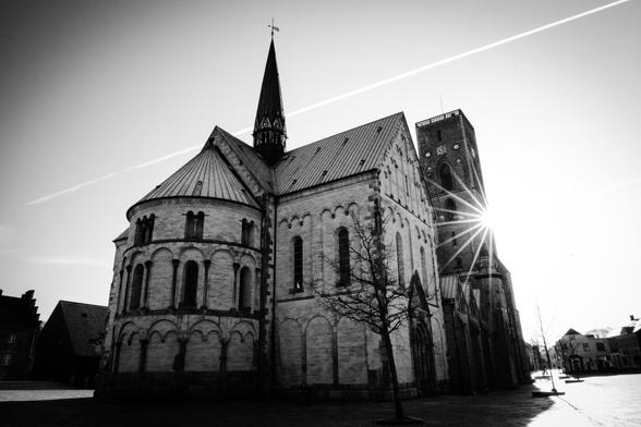 Scwarzweißaufnahme des Doms von Ribe mit einem Sonnenstern, der hinter einem hohen eckigen Turm hervorblitzt. Hinter dem Dom führt ein Kondensstreifen diagonal und ca parallel zur Dachkante von rechts oben nach links mittig.