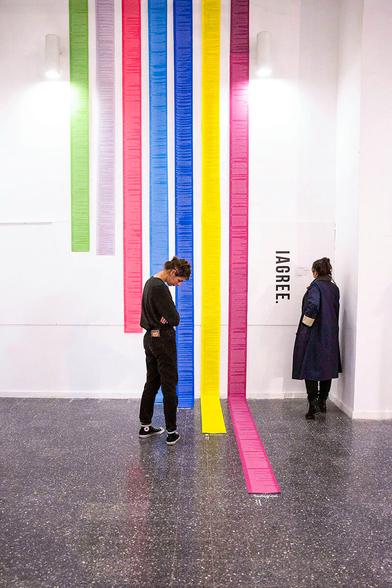 Alt-Txt: Two people stand before multi-color strips of paper with text on them that stretch down an entire wall and onto the floor. The title of the installation "I Agree" appear in all caps on the wall, too.