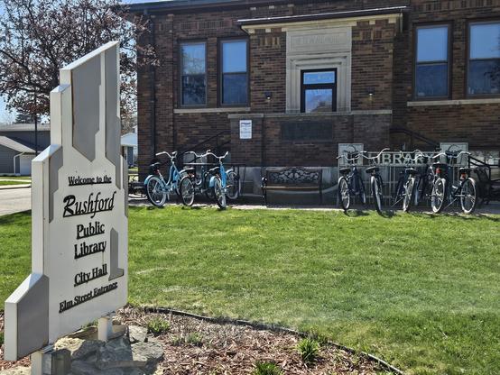 A white and grey sign says "Welcome to Rushford Public Library. City Hall. Elm Street Entrance. The grass is green. 4 blue bicycles in a bicycle parking rack. Above it is a white signthat says "Bike Share of Rushford". 5 black or grey bicycles in another bicycle parking rack that has the word "Library" formed in it. A building with a brick wall with 4 windows and a glass door in the middle.
