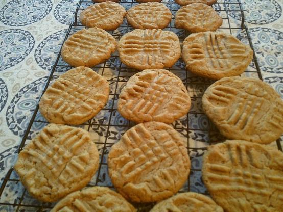 Several golden-brown peanut butter cookies are arranged on a black wire cooling rack. Each cookie has the characteristic criss-cross fork marks on top. The cookies vary slightly in shape and size.
