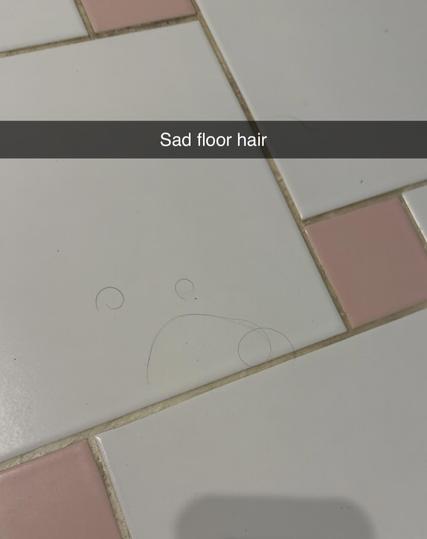 Some black hairs on some old-style bathroom floor tiles. They form a kind of frowny face; two small curled up ones (body hair; they are not pubes, I promise!) being the eyes and a longer one being the mouth.