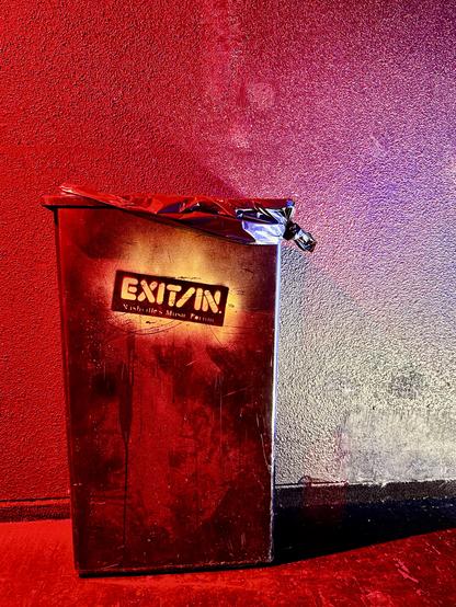 A rectangular trashcan with the words exit/in written on it and spray paint stencil. Bathed in red light and looking pretty worn and wise.