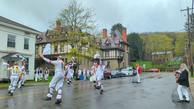 Six morris dancers with white vests, shirts, and trousers, decorated with red, blue, and yellow ribbons, are dancing around a circle in a street that is wet from recent rain. To their right someone is making a video, and on the far right are two musicians playing melodeons. Behind them are more morris dancers, some dressed similarly and some in black vests, watching. At the back is a large Tudor revival inn.