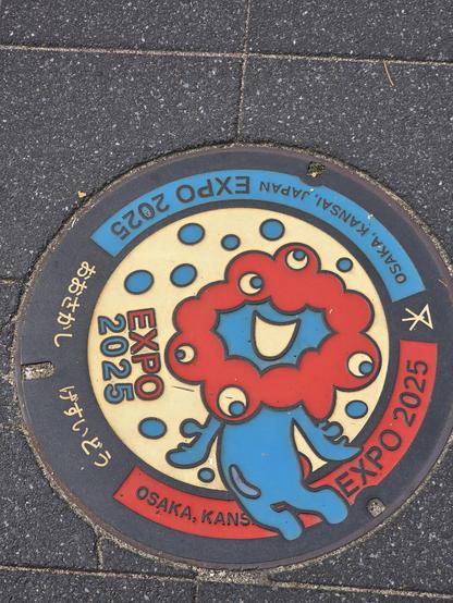 A manhole cover depicting the eldritch horror that is the Osaka Expo 2025 monstrosity. This cover is colorful with blue and red and light yellow