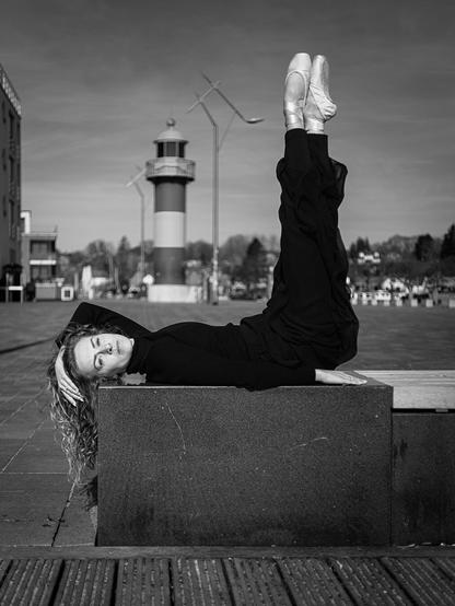Ballettänzerin liegt mit ihrem Rücken auf einem Steinquader und streckt ihre Beine in Spitze hoch. Hinter ihr sieht man in der Unschärfe den kleinen Leuchtturm an der Hafenspitze von Eckernförde.
