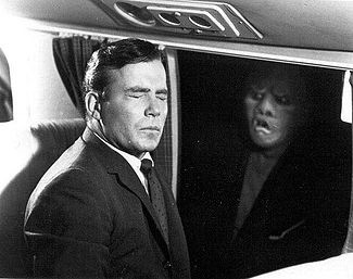 A black and white still of William Shatner sitting in an airplane seat next to the window. His eyes are closed. A humanoid creature with evil eyes and a slanted mouth is looking at him from outside.