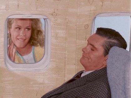 A still from Bewitched of Samantha looking at her sleeping husband from the window of an airplane. She is flying outside and following the plane. She has a playful smile.
