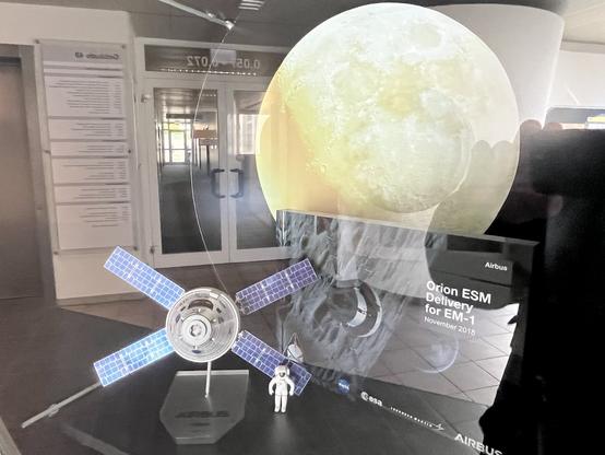 The image features a display showcasing a model of the Orion spacecraft's European Service Module (ESM) along with a small astronaut figurine. In the background, there's an artistic representation of the Moon. The display is labeled "Orion ESM Delivery