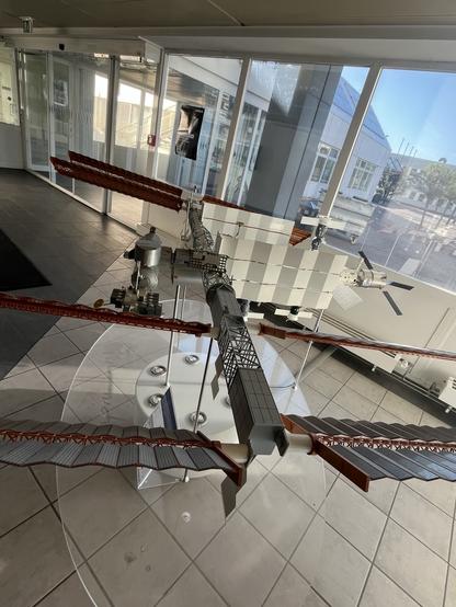 A detailed model of a space station is displayed on a circular glass table inside a modern building. The model features solar panels, docking ports, and various scientific instruments, highlighting its intricate design. Large windows provide a view of the outside environment.