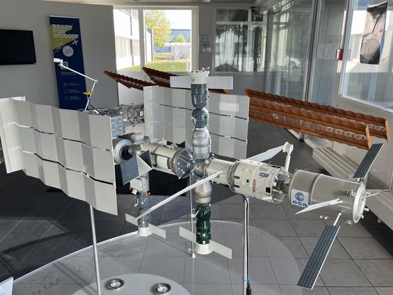 A model of the International Space Station (ISS) is displayed indoors, featuring detailed components like solar panels, modules, and antennas. The background includes informational signage and a partially visible structure.