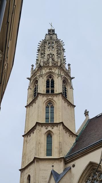 Das Bild zeigt die majestätische Spitze einer gotischen Kirchturmspitze, die in den Himmel ragt. Die Turmspitze ist mit komplexen Steinreliefs und Skulpturen verziert, die typisch für die gotische Architektur sind. Die Fenster sind schmal und hoch, mit gotischen Bögen, die das Licht durch lassen. Die Fassade des Turms ist aus hellem Stein gefertigt und zeigt Spuren der Zeit. Im Hintergrund ist ein Teil eines anderen Gebäudes zu sehen, das ebenfalls gotische Elemente aufweist. Der Himmel ist bewölkt, was dem Bild eine ruhige und historische Atmosphäre verleiht.