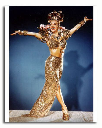 A color photograph of Carmen Miranda in a long, tight, golden two-piece dress, accents, and a head wrap. She stands in pageantry, with her arms outstretched and a broad smile, against a blue background.