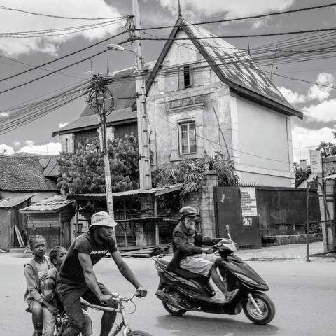 Un scooter à barbe doublant un vélo-transport-familial devant une maison ancienne typique du quartier (dans le style d'avant le bétonnage général ayant court actuellement)