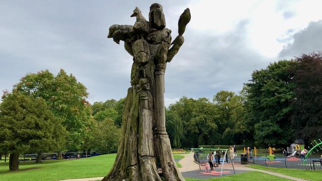 This picture shows a large, intricately carved wooden sculpture standing in a park. The sculpture appears to be made from a tree trunk, with various detailed carvings of animals and human figures. Among the carvings, you can see birds, and other creatures, as well as faces and figures of people. The sculpture stands tall with weathered textures adding to its character.

In the background, there is a playground with children and adults enjoying the play equipment. The park is lush and green, with large trees surrounding the area. The sky is overcast, giving a slightly grayish hue to the scene. Paved pathways and manicured grass add to the well-maintained park environment.
