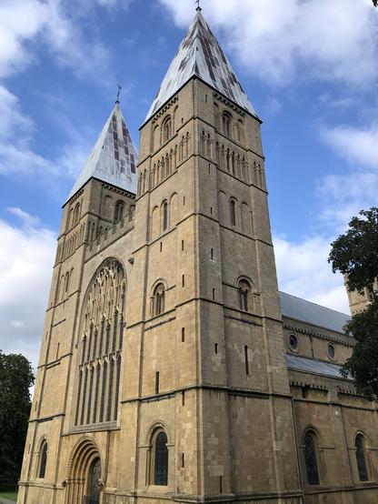 The photograph is taken from the south west of the exterior of the magnificent west end with twin towers and many Romanesque features such as round-arched doorway and windows. Parts of the south elevation of the nave are visible with aisle and clerestory. The light honey coloured stone glows in the sunshine against a blue and white summer sky. Photo copyright Coralie Mills 2023
