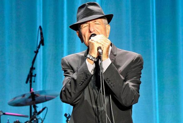 Leonard Cohen, vêtu d'un costume sombre et d'un chapeau noir, interprète une chanson au micro sur la scène du King’s Garden à Odense, Danemark, en 2013. En arrière-plan, on distingue une batterie et des équipements de scène.