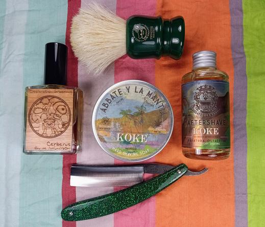 (Le Chat Mistral description)

This image shows a collection of men's grooming items arranged on a colourful striped cloth. There's a wooden bottle labelled "Cerberus" with a circular design, likely a cologne. Next to it is a round tin of "Koke" hard shaving soap, featuring a tropical scene. A matching "Koke" aftershave bottle is nearby. A green shaving brush with a dark handle is placed above these items. At the bottom, there's a straight razor with a green glittery handle. The items are neatly displayed, suggesting a focus on traditional shaving and grooming.