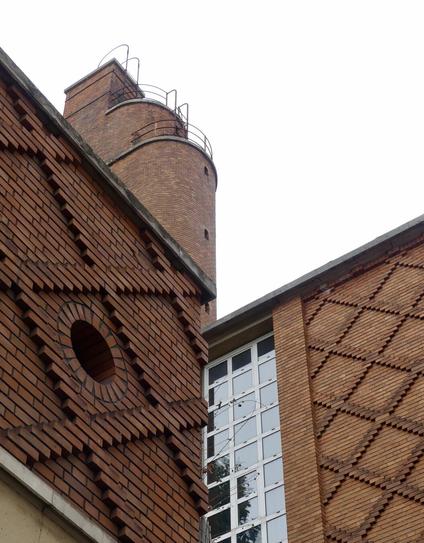 Vue en contreplongée de l’angle où se rejoignent deux parties d’un bâtiment aux lignes géométriques sobres années 30. Au premier plan, les murs extérieurs sont recouverts de croisillons en briques, à l’exception d’une partie en verre qui doit correspondre à l’escalier à l’intérieur. Un peu plus haut, on devine une partie surélevée aux lignes arrondies toujours en briques, mais sans motifs. Le ciel est gris, la lumière pas top.
