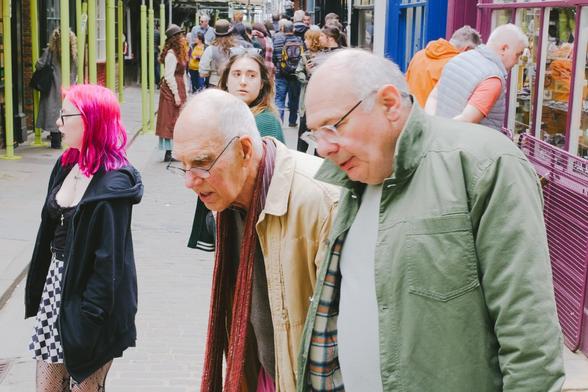 This vibrant and bustling street scene captures a diverse group of people walking along a colorful sidewalk. In the foreground, two older men are engaged in conversation. The man on the left wears a beige jacket and glasses, while the man on the right is dressed in a green jacket and also wears glasses. To their left, a person with bright pink hair stands out, wearing a black jacket and a black-and-white checkered skirt, adding a splash of color to the scene.
Behind them, the sidewalk is lined with people dressed in various styles, including some in historical or period costumes, suggesting a festive or themed event. The background features colorful storefronts with yellow, blue, and purple facades, creating a lively and energetic atmosphere. The overall mood is one of community and celebration, with people of all ages and styles coming together in this vibrant setting.