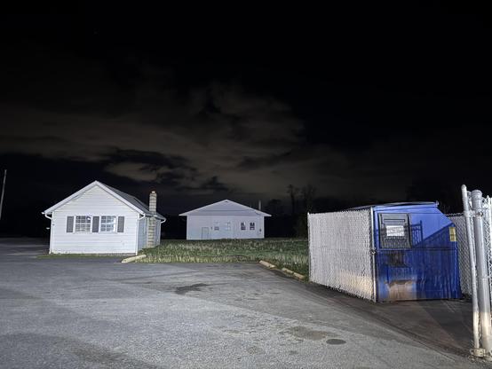 Two small single story buildings painted white sit at the edge of a parking lot. A fenced in blue dumpster is here as well. It is late at night. No lights or life in the local hamlet. It was chilly, quiet, and scary when I took this. I didn’t stay long.