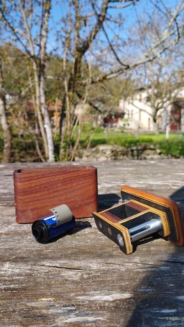 Stenope ouvert en 2, posé sur une table de pique-nique en bois, une pellicule enroulée dans son boîtier posée à côté, l'amorce dépassant. En arrière plan, une rivière, des arbres et une maison. Le ciel ert bleu. L'ambiance bucolique