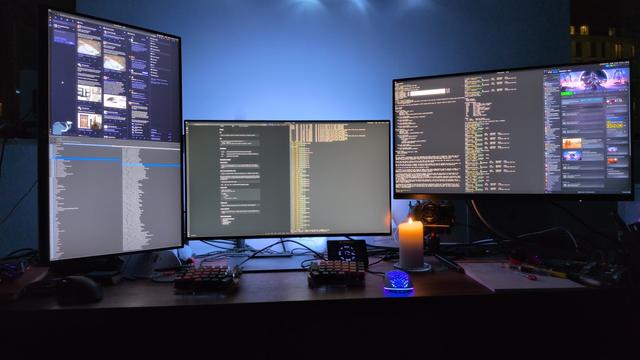 A picture of my desktop at night.  Three 27" monitors are arrayed on a wide desk.  The leftmost is aligned vertically, the other two horizontally, with the rightmost a few inches higher than the centre one.  The desk has a split keyboard in the centre and a white mouse to the right, glowing indigo.  A candle is lit near the mouse next to a large A4 pad.  Various pens are spread around, and a Stream Deck and Wacom pad.  The left monitor has Mastodon open in a web browser on its top half and a music player in its bottom half.  The centre browser has two pages open, one a dark page of documentation for something or other, the other a dark terminal.  The rightmost monitor had three windows open, two terminals and one Steam window.  One of the terminals has a code example printed.