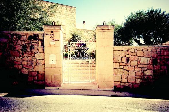 Schmiedeeiserne weiß gestrichene Eingangstür eines sonnenbeschienenen Hauses in einem Ort auf Malta mit der treffenden Bezeichnung 'Paradis du soleil' auf dem Steinpfosten links. Steinmauer links und rechts, dahinter sichtbar ein Teil des Hofes und Hauses aus hellem Stein und grüne Bäume.
