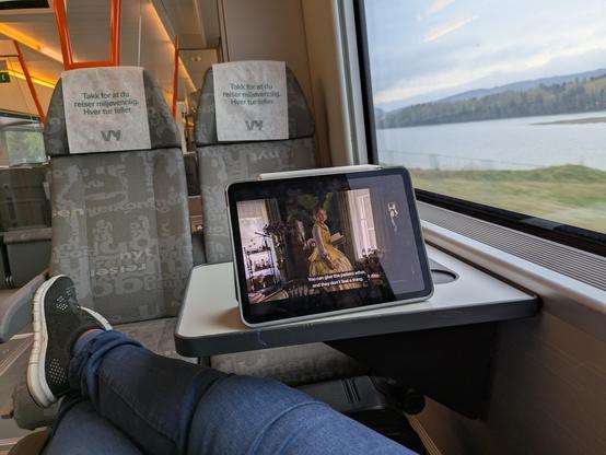Picture showing POV of a train passenger, gray seats. Legs resting on a suitcase and an iPad showing a scene from the artful dodger.