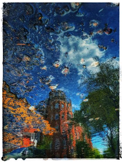 Das Bild zeigt eine Reflexion einer städtischen Szene in einer Wasserpfütze. Zu sehen ist die Spiegelung eines roten Backsteinturms, der von grünen Bäumen umgeben ist. Darüber breitet sich ein blauer Himmel mit weißen Wolken aus. Die Reflexion im Wasser ist leicht verzerrt durch kleine Bewegungen des Wassers, was dem Bild eine impressionistische, fast malerische Wirkung verleiht. Das Foto ist um 180 Grad gedreht.