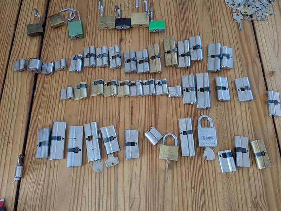 Das Bild zeigt eine große Anzahl von Zylinderschlössern und Vorhängeschlössern, die ordentlich auf einer Holzoberfläche ausgebreitet sind.
Die meisten Schlösser sind Zylinderkernschlösser aus Metall (größtenteils silbern, einige messingfarben), viele davon sind Doppelzylinder, einige auch Halbzylinder.
Oben im Bild befinden sich mehrere Vorhängeschlösser, darunter einige mit grünem oder messingfarbenem Gehäuse.
In der rechten oberen Ecke liegt ein Haufen verschiedener Schlüssel lose auf dem Tisch.
Weiter unten im Bild sieht man ein paar ABUS-Schlösser, darunter ein großes silbernes Vorhängeschloss mit der Aufschrift "ABUS TITANIUM".
Die Schlösser sind in mehreren Reihen angeordnet, teils aufrecht, teils liegend.
Es scheint sich um eine Sammlung oder Vorbereitung für Reparatur, Sortierung oder Training (z.B. Lockpicking) zu handeln.