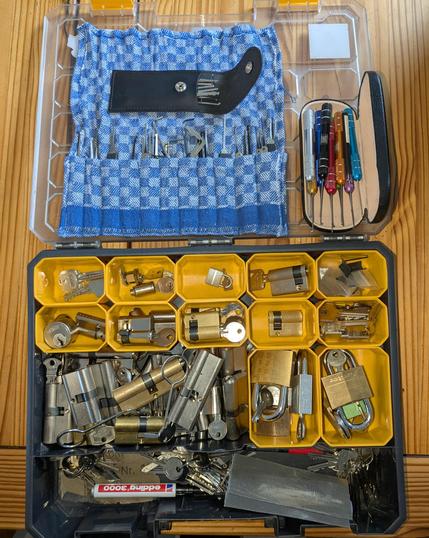 Das Bild zeigt eine offene Werkzeugkiste auf einem Holztisch.
In der oberen Hälfte der Kiste befindet sich ein blau-weiß kariertes Tuch, in das zahlreiche Lockpicking-Werkzeuge ordentlich eingesteckt sind. Rechts daneben sind mehrere bunte Spezialwerkzeuge in einer kleinen schwarzen Hülle verstaut – vermutlich Pinzette oder Feilen für Feinmechanikarbeiten.
Die untere Hälfte der Kiste ist in viele kleine gelbe Fächer unterteilt, in denen verschiedene Schließzylinder, Vorhängeschlösser und Schlüssel sortiert sind.
Links unten liegt ein Bereich mit vielen Schlüsseln, einem Edding-Stift (Modell 3000) und weiteren Kleinteilen.
Man erkennt sowohl Messing- als auch silberfarbene Zylinder und Vorhängeschlösser, teils von bekannten Marken wie ABUS.
Die ganze Ausrüstung wirkt sehr strukturiert und organisiert – sie scheint für Lockpicking, Schließtechnik-Training oder Reparaturarbeiten gedacht zu sein.