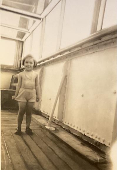 A sepia photo of a small girl in a summer romper, standing on the inside deck of a ship in 1946. She looks quite pleased with herself.